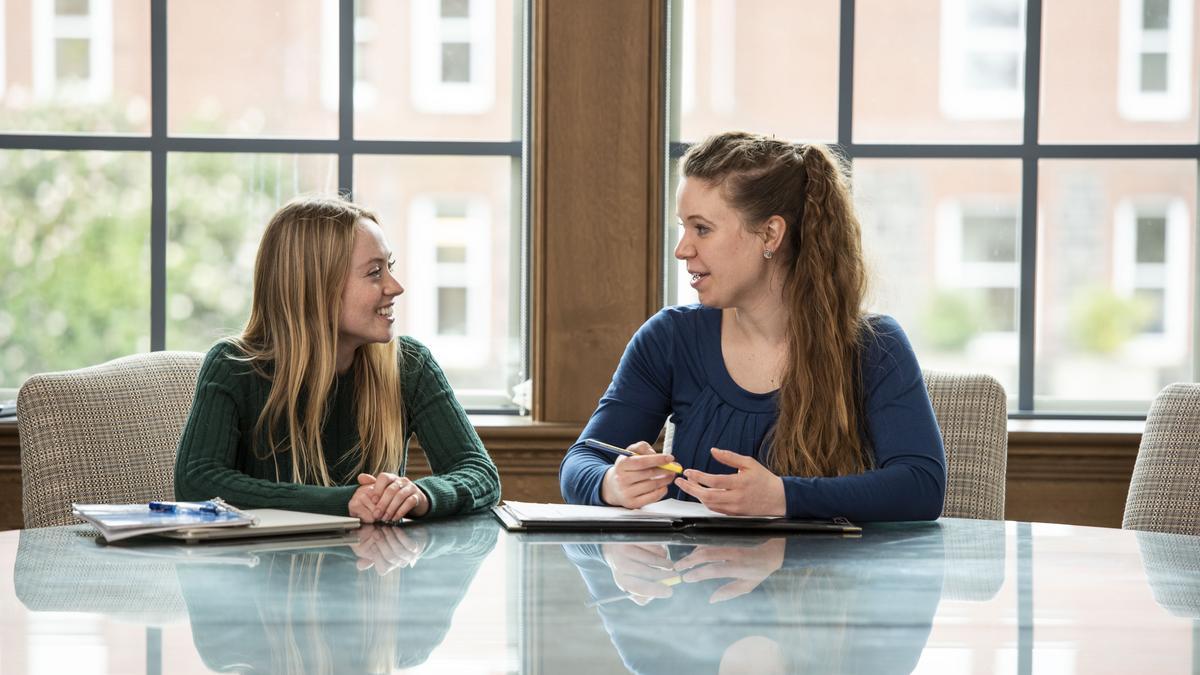 female student sitting at glass table talking with female staff member with windows in background