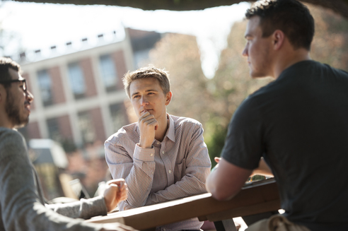 Three young men sit around outdoor table under tree talking