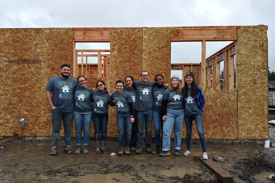 A volunteer group poses for a picture infront of a framed out house in the process of being built.