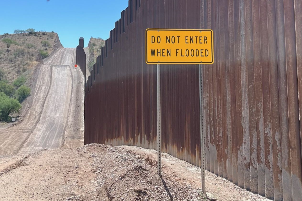 A high steel border wall lines one side of a dusty road with a sign posted that states "Do not enter when flooded"