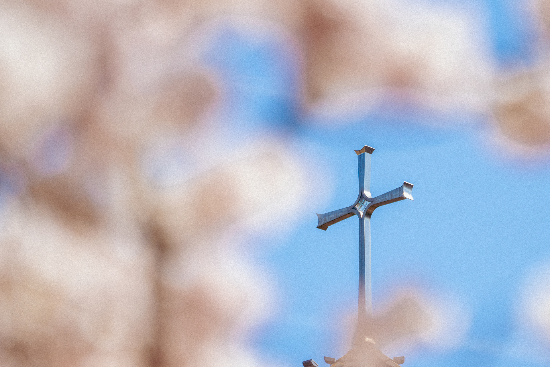 A close-up of a silver cross steeple atop a chapel bell tower framed by a blurry pink cherry blossom tree.