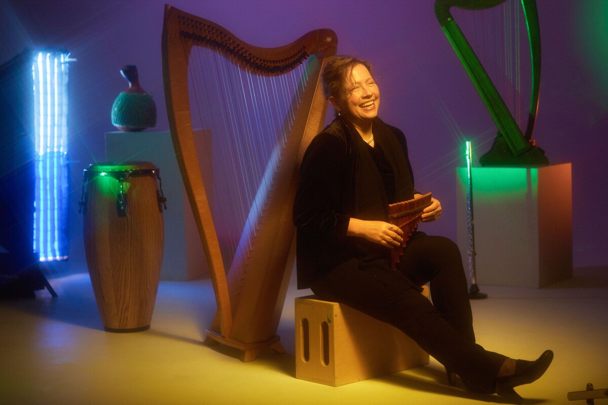 Mo Briare sits on a bench in front of a harp, holding a pan flute and smiling.