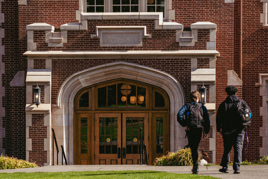 A behind view of two students wearing backpacks walking toward the arched entrance of Dundon Berchtold Hall.