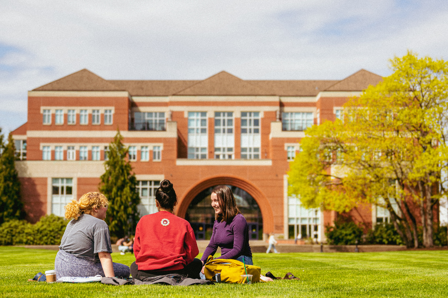Three students sit on the grass together in front of the Franz Center during the summer, smiling and talking. 