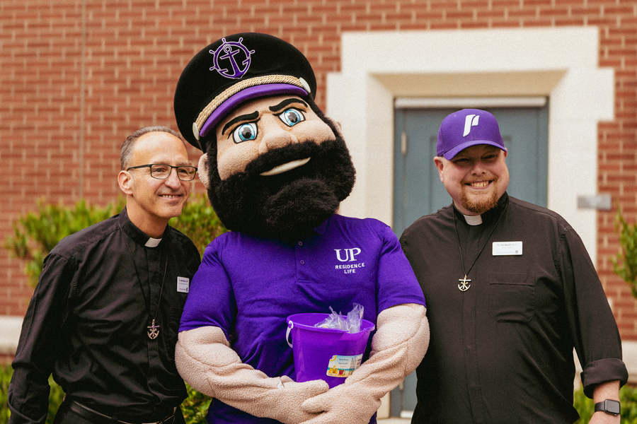 Two priests wearing clerical collars stand and smile with University of Portland's mascot, Wally the Pilot.