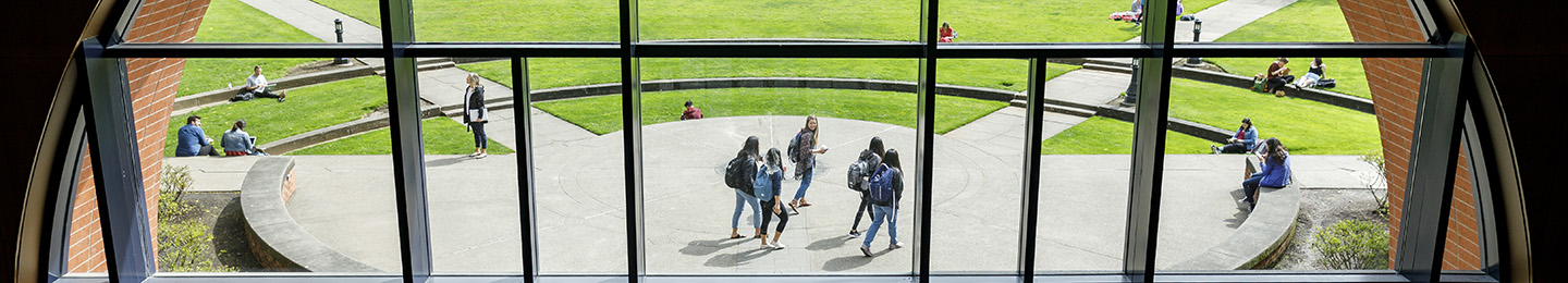A view through a large window showing students walking on a pathway toward the University of Portland Quad