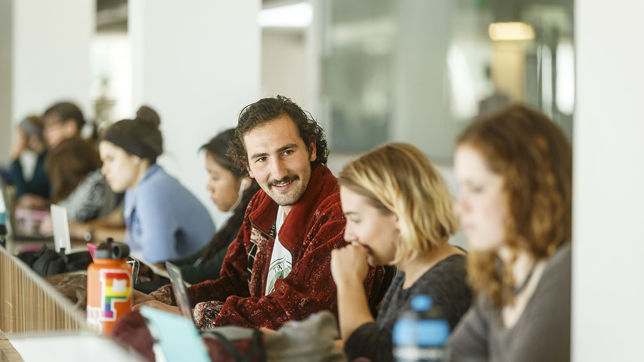 A student with a mustache looks over, smiling at his peers in a classroom