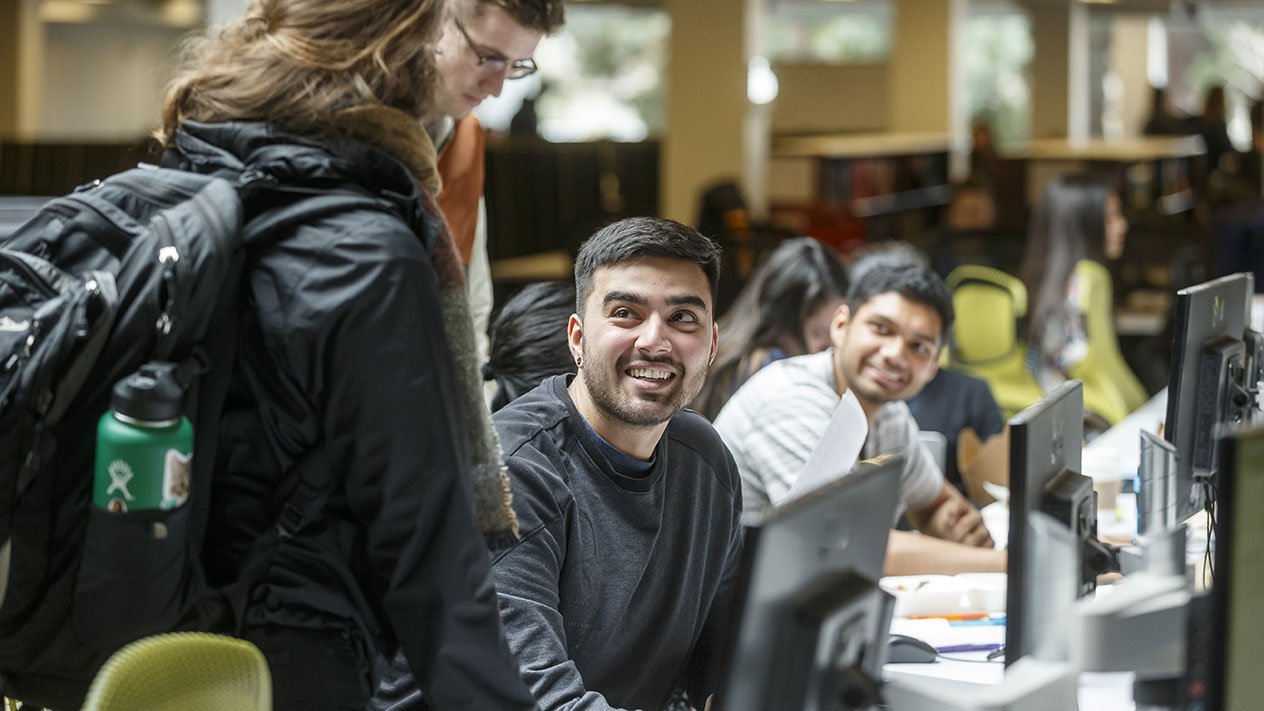 Students sitting at a row of computers in the Clark Library, smiling up at a standing student