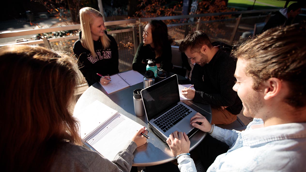 A group of friends sit at a table outside, one student looking at their laptop screen