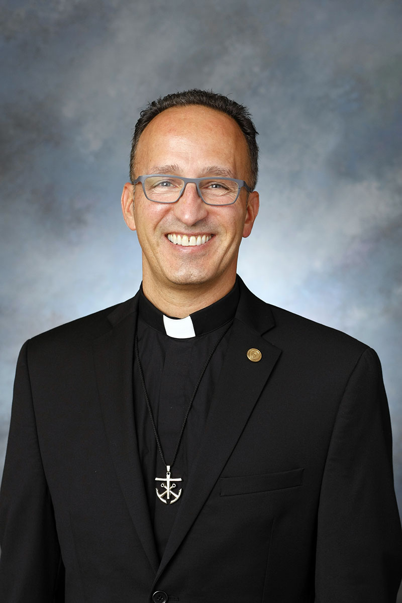 Father John Donato against a blue portrait backdrop, smiling.