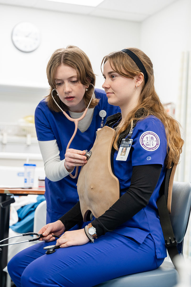 two nursing students practice listening to chest sounds on a medical manikin in the simulation lab