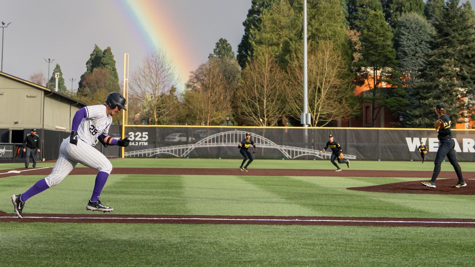 A Pilots baseball player hits the ball while at bat during a game with a rainbow in the sky behind the field.
