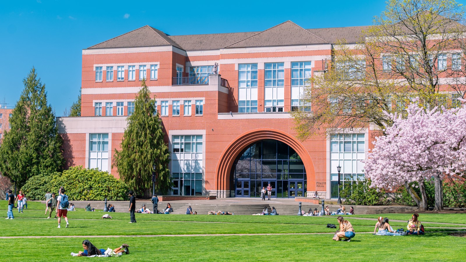 The exterior of Franz Hall, a modern brick building, on a sunny day. The grassy quad is lined with students and blossoming trees.