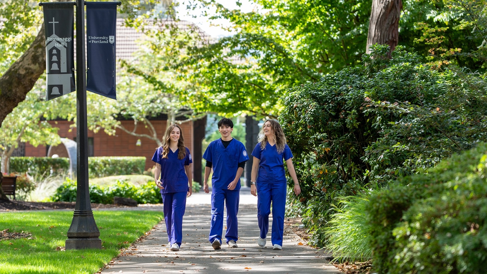 Three UP nursing students in scrubs walk along a campus path on a sunny day.