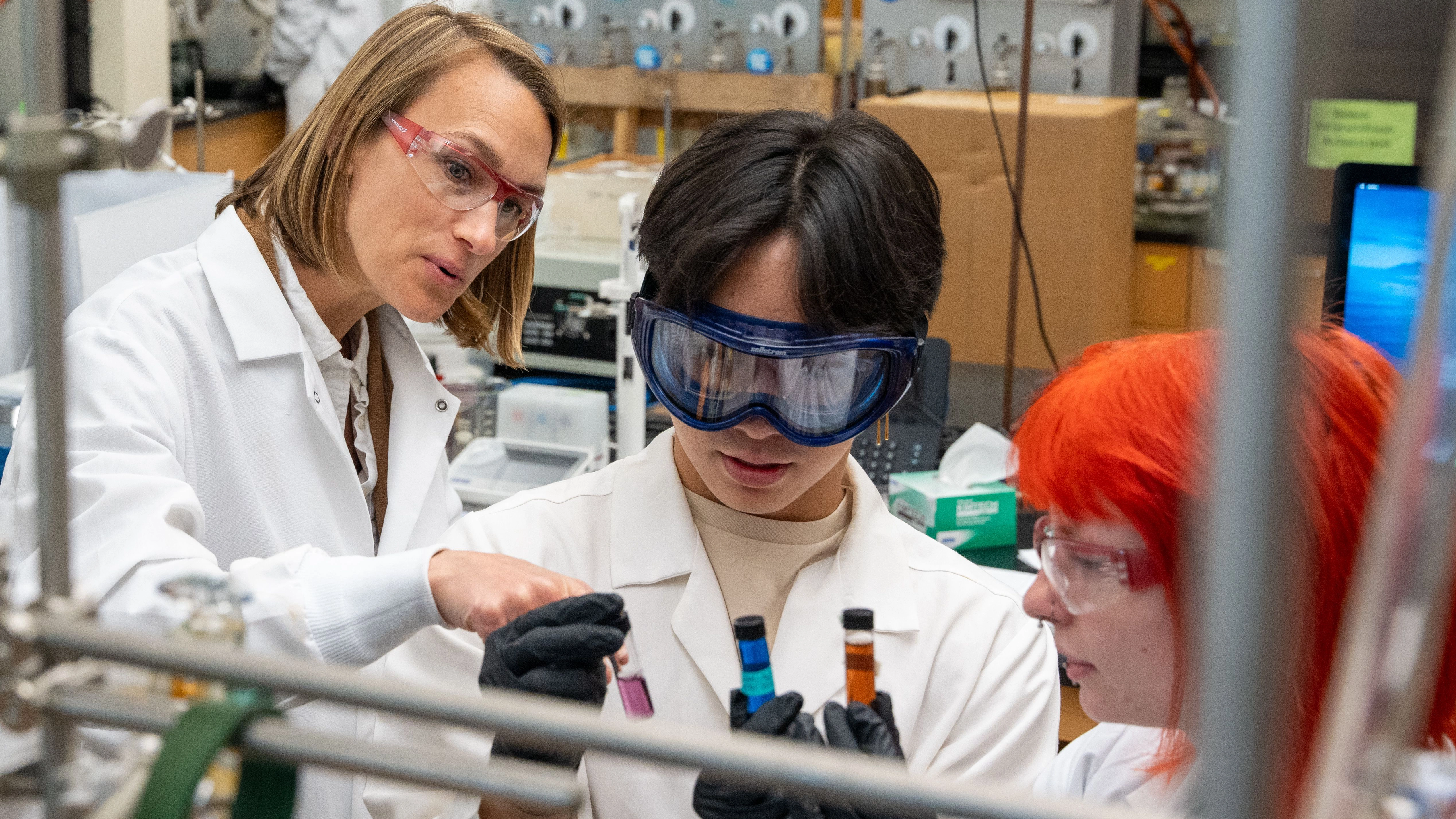 A teacher stands with two students in a lab setting