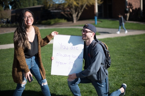 boy on his knee holding sign that reads. "you are always look great 25/8" girl laughing