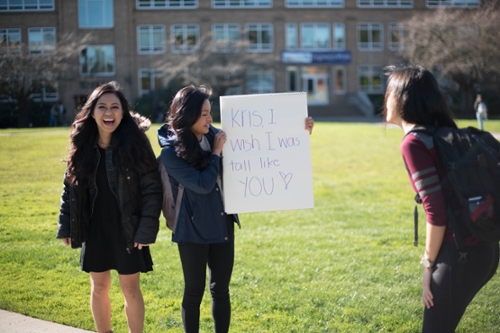 three girls laughing, one girl reading sign that reads, "Kris, I wish I was tall like you!"