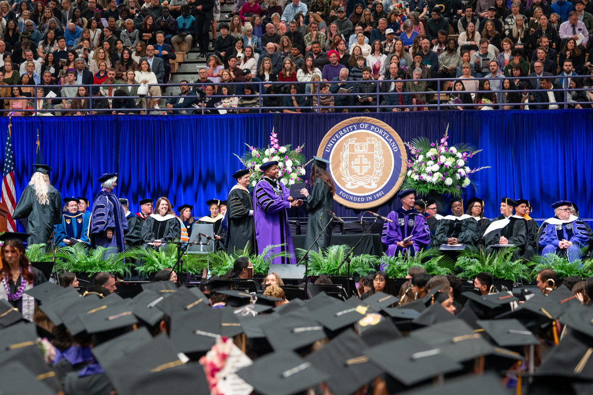 A crowd of seated graduates in the foreground watches as students receive their diplomas on the stage, with spectators seated in stands behind them.