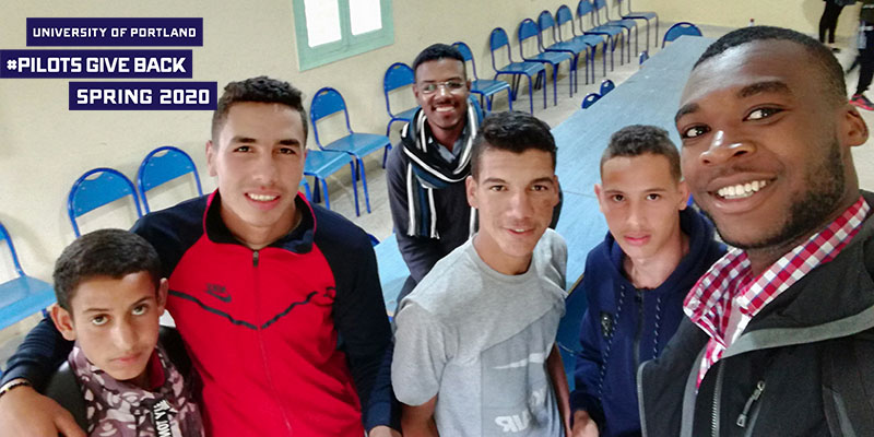 Kene Angibogu with friends from Morocco smiling with logo that reads University of Portland Pilots Give Back Spring 2020