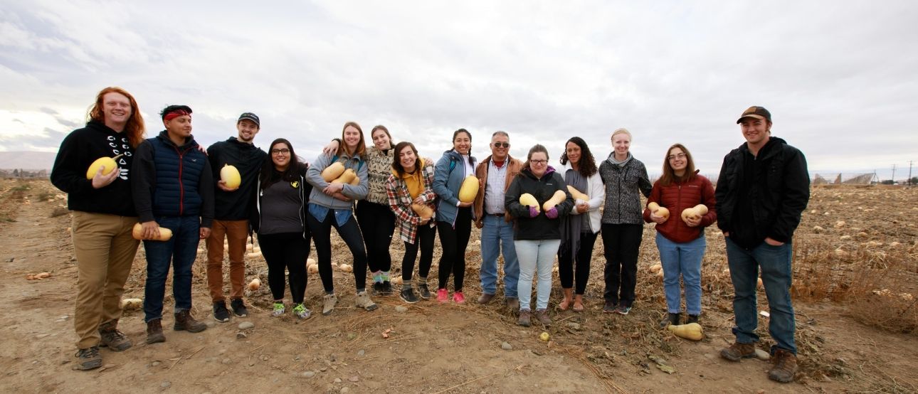 A friendly group of people standing in an agricultural field smile into the camera while holding winter squash in their hands.