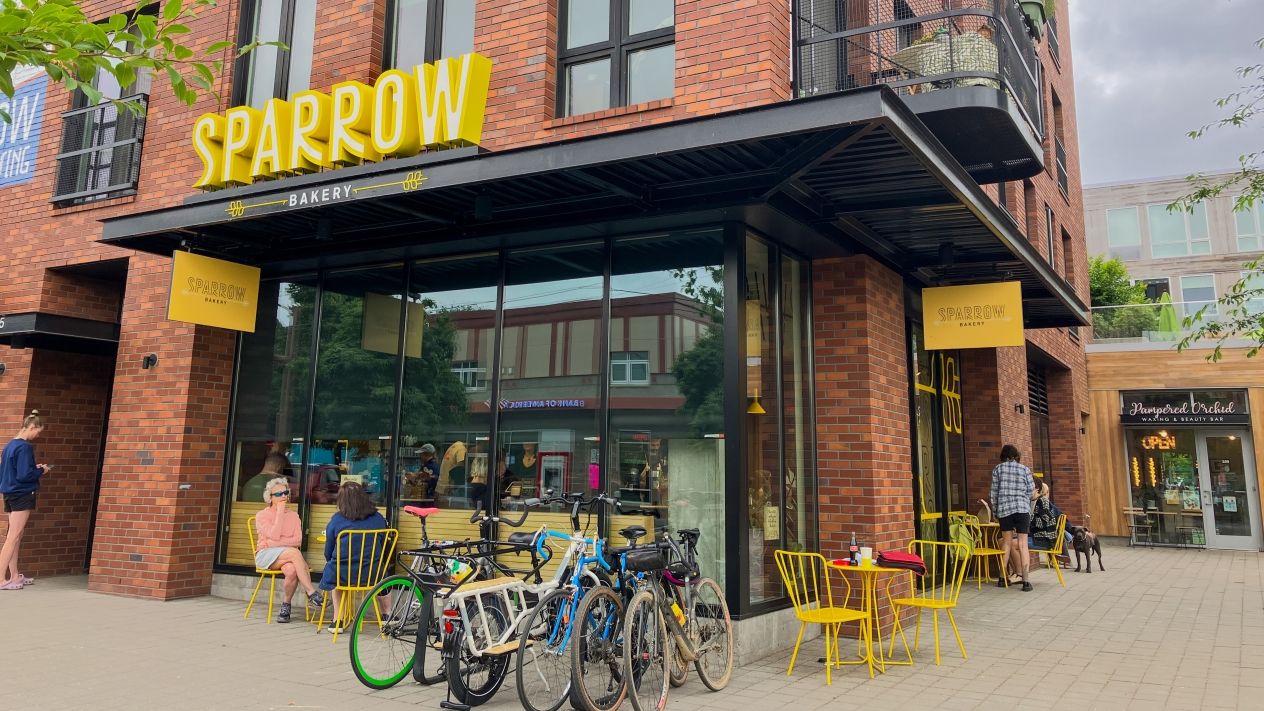The facade of Sparrow Bakery, a modern brick building with patrons gathered outside at cafe tables.