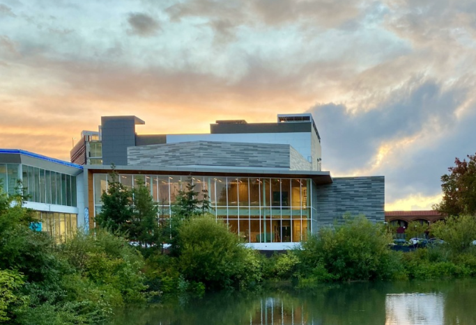 Exterior of the Reser Theater at sunset, a contemporary building with large windows sitting at the edge of a pond that is fringed with vegetation.