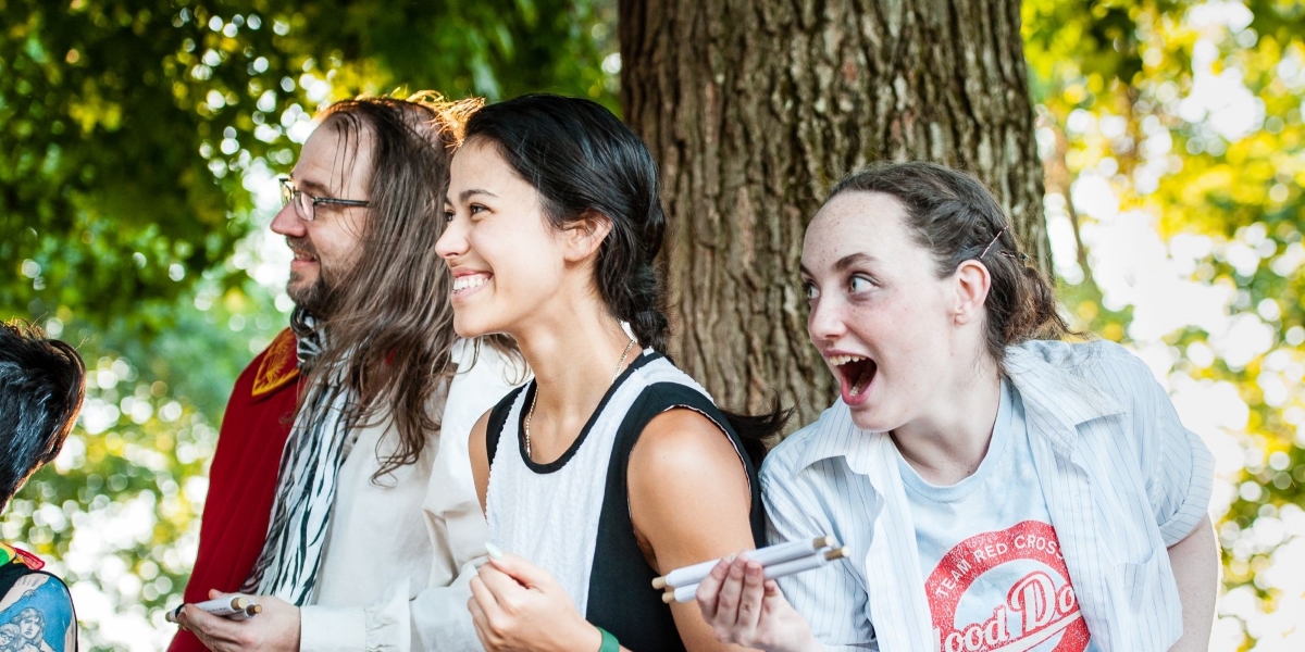 Actors with expressions of surprised delight stand under a tree on a sunny day in the park during a performance.