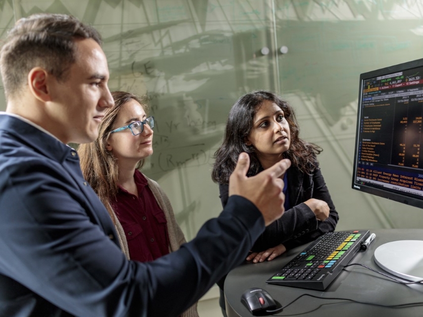 Three people are gathered around a trading terminal looking intently at the data on the screen.
