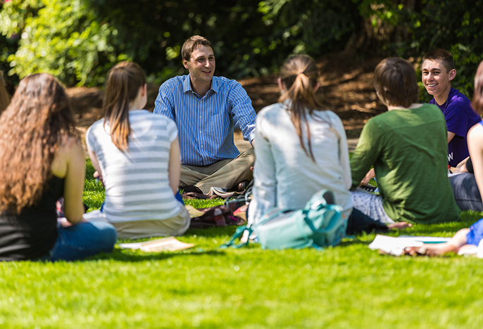 Teacher and students sitting in a circle on the grass having class outside.