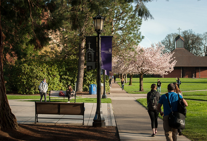 students walking on pathway on campus on a sunny day. Lamppost with purple UP sign in foreground. 