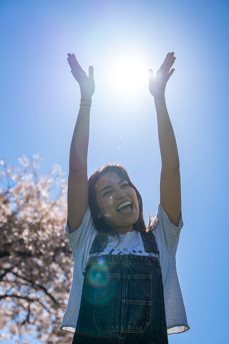 smiling student frames sun with hands in front of cherry blossom trees