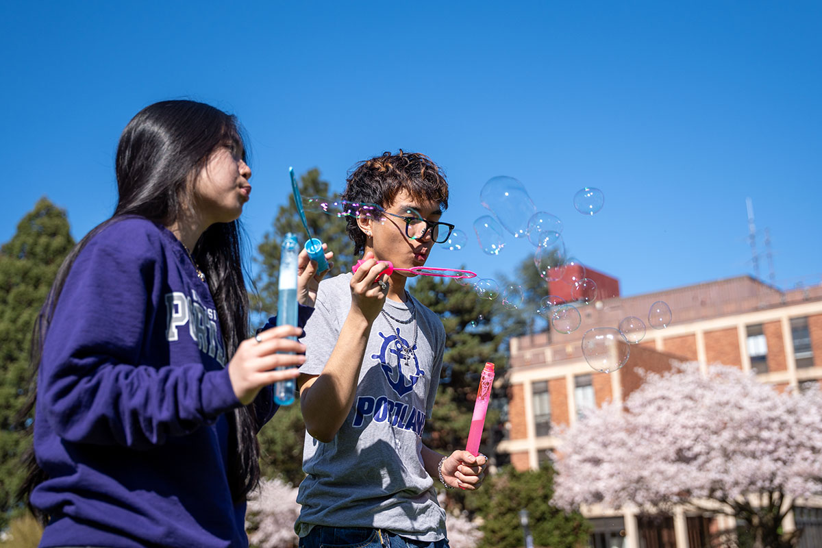 students blowing bubbles from bubble wands on the quad with blue sky and cherry blossom trees behind them
