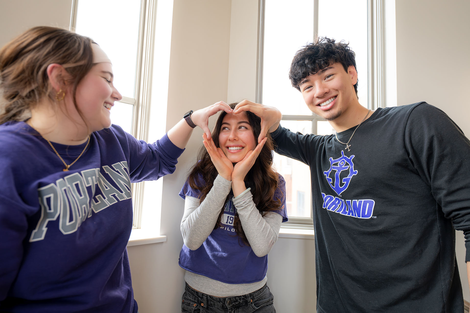 three students making a heart shape with their hands around the smiling face of the middle person