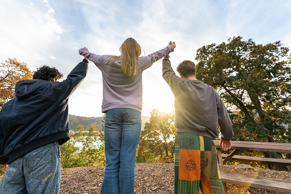 Two students help uplift a third student between them with golden sun shining behind them off the river