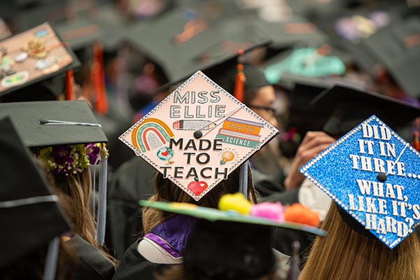 decorated mortarboards at commencement, one that says 'Miss Ellie made to teach' and another that says 'did it in three... what, like its hard?'
