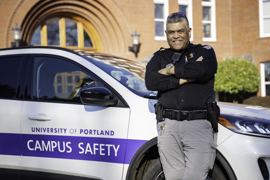 Officer Reyn Catala stands with his arms crossed in front of a University of Portland campus safety vehicle.