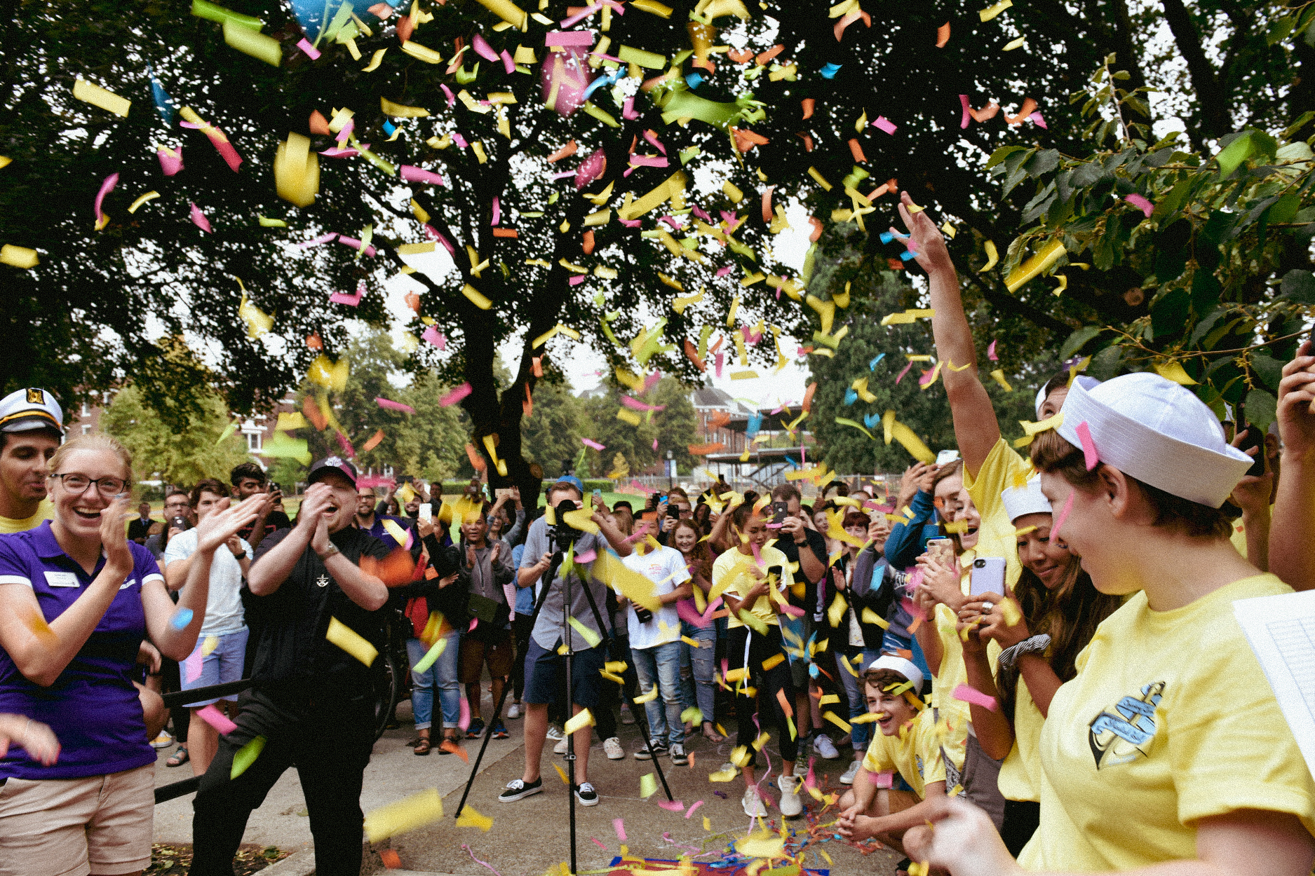 A large crowd of students and staff stands together under a shower of colorful confetti on move-in day.
