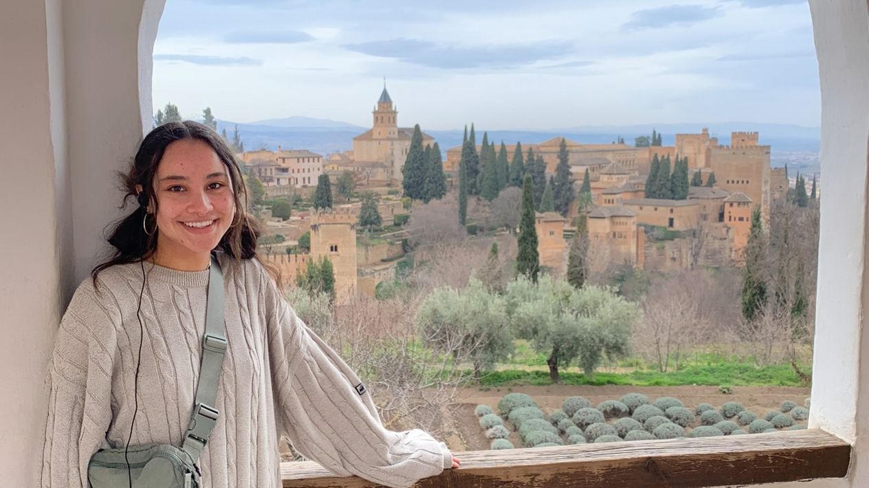 A study abroad student stands by a window that looks out onto a picturesque view of a European village.