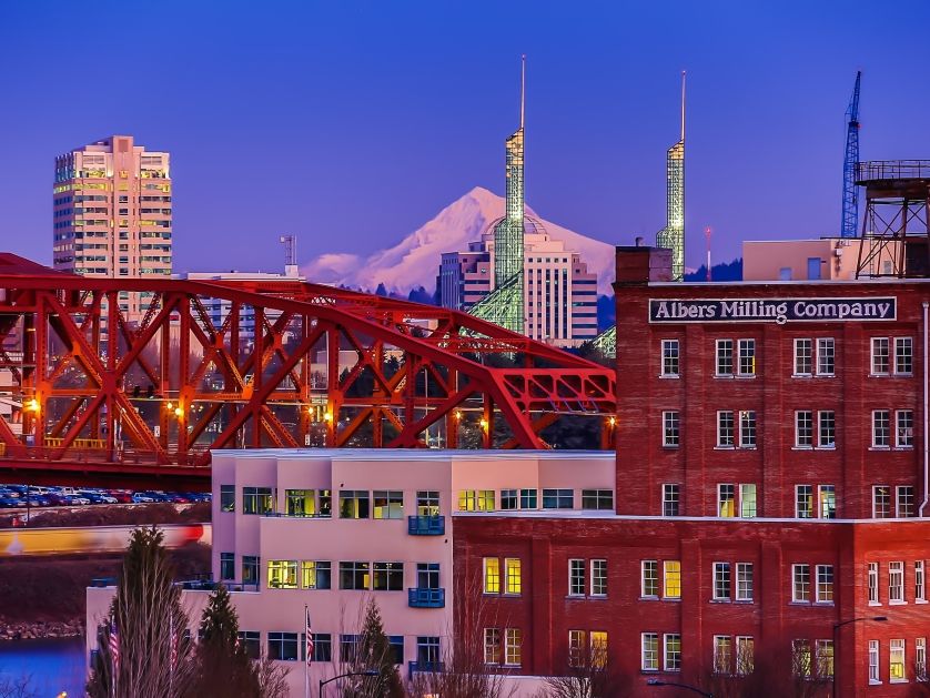 The Portland skyline at dusk with bridges, factory buildings, and Mt. Hood in the background.