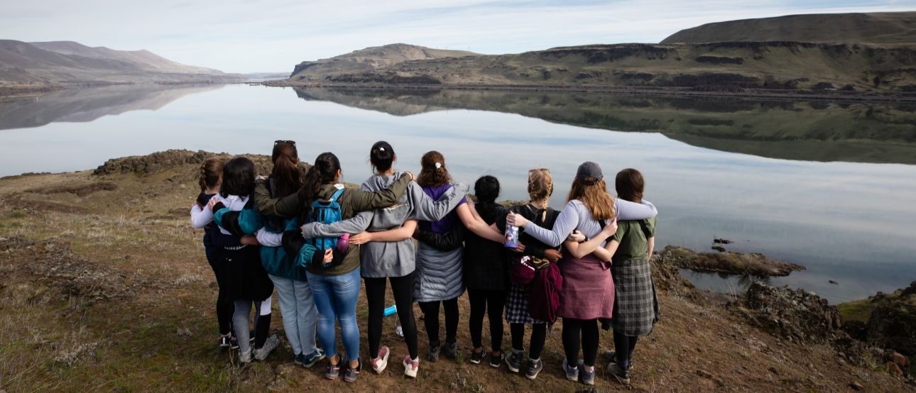 A group of students seen from behind with their arms linked looking into a scenic river valley.